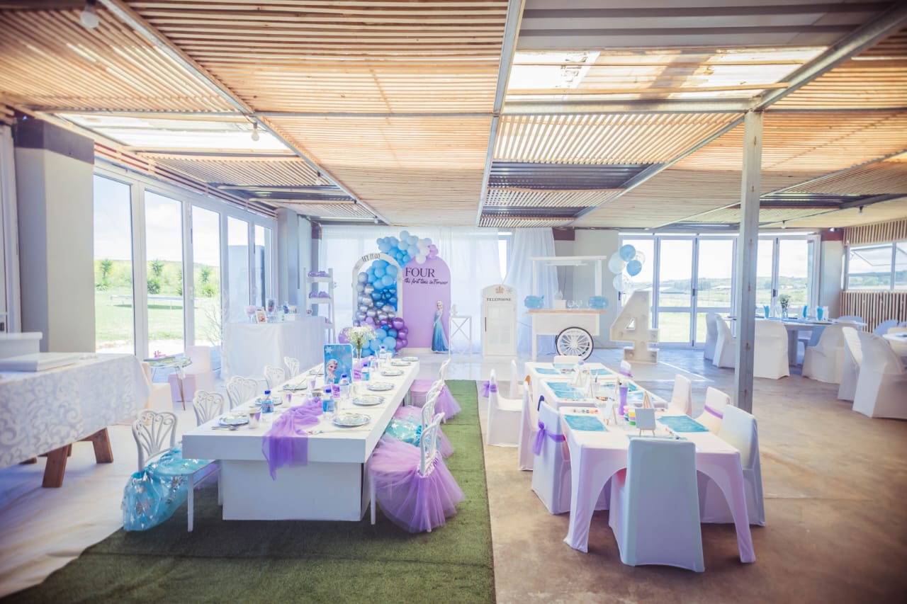 The Mini & Me party hall set up for a Frozen-themed birthday with low kids’ tables, a dessert cart and a light-up number four