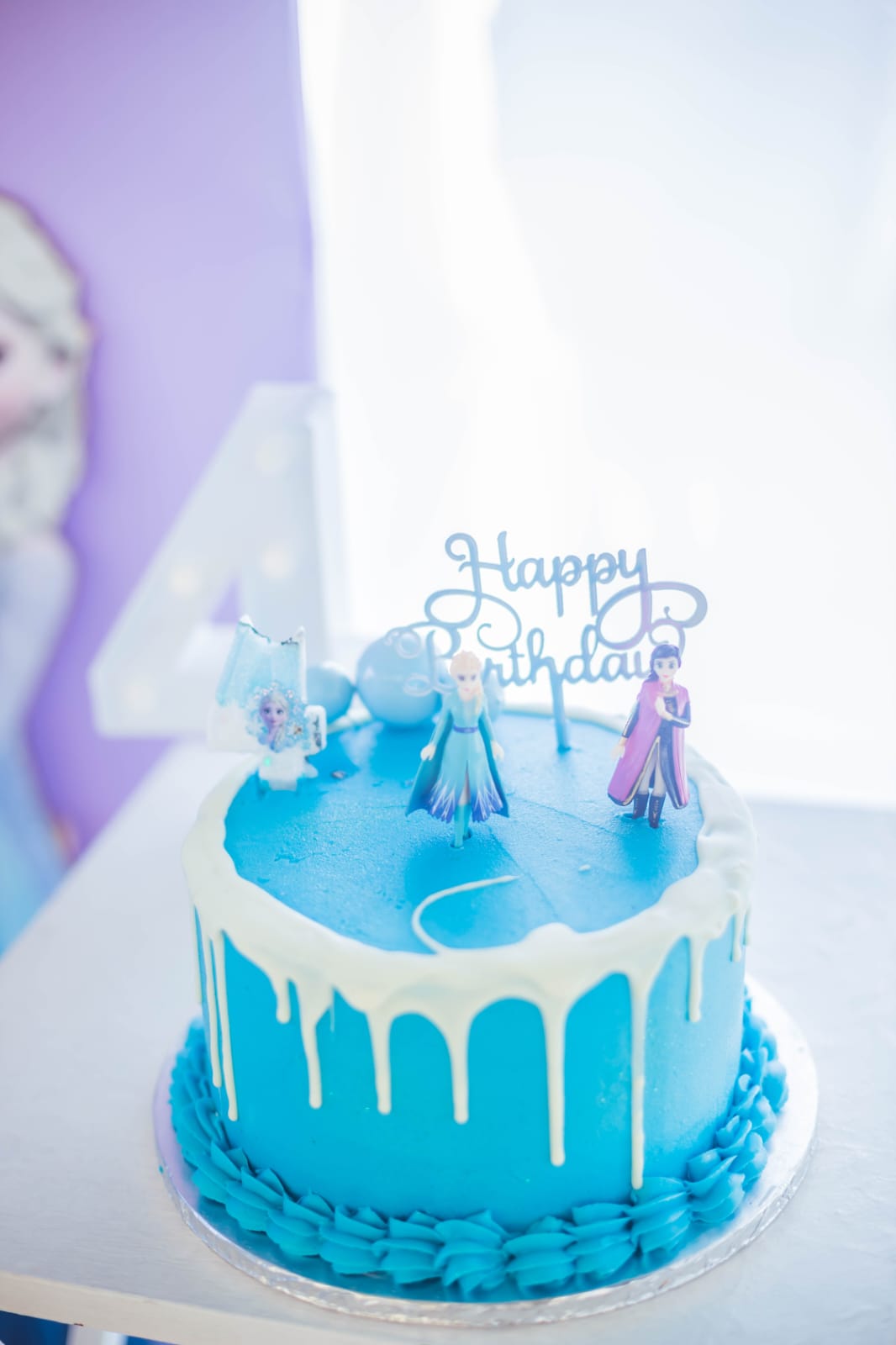 Frozen birthday cake with Elsa and Anna toppers and a Happy Birthday sign, in front of a lilac FOUR backdrop