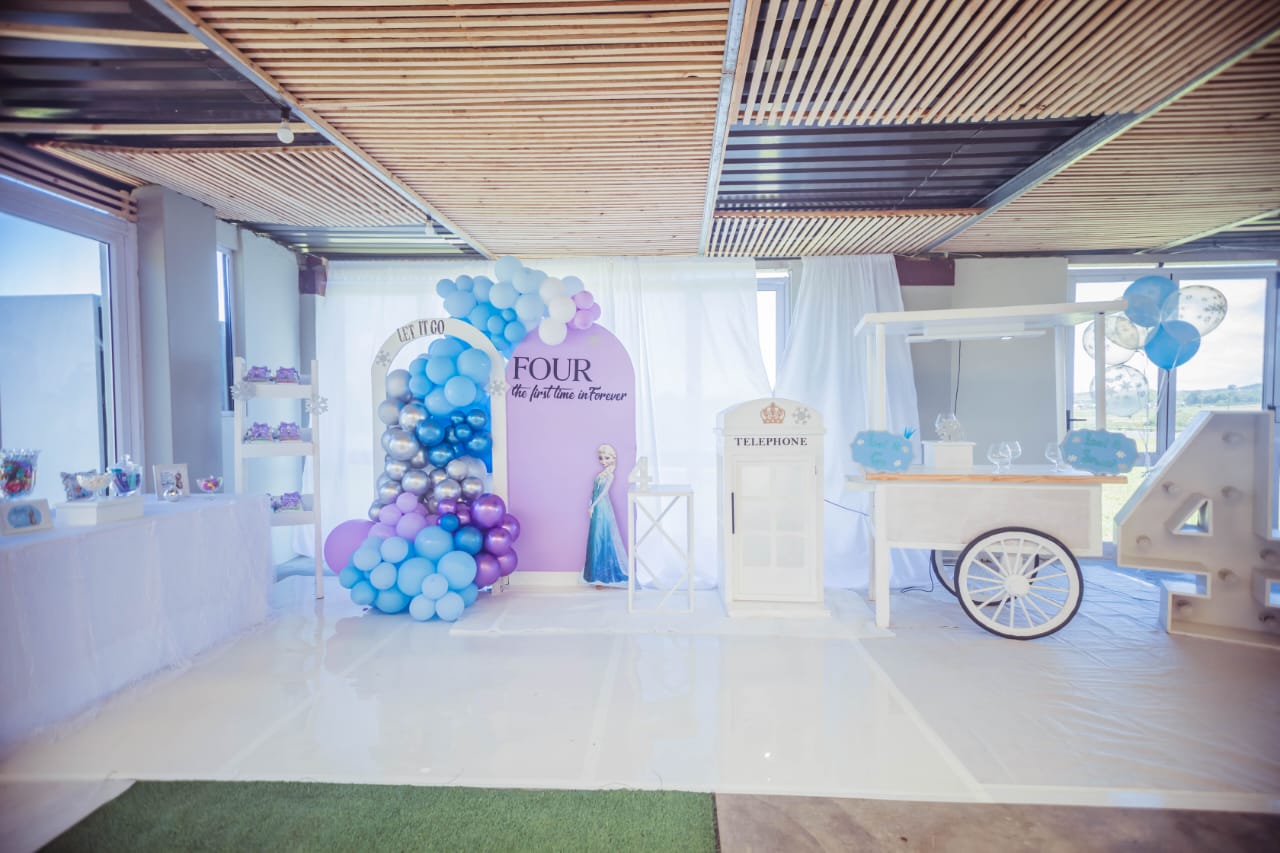 Frozen party décor with a “Let It Go” arch framed by a blue and silver balloon garland, a lavender Elsa backdrop, a white telephone booth, a sweet cart and an illuminated number 4